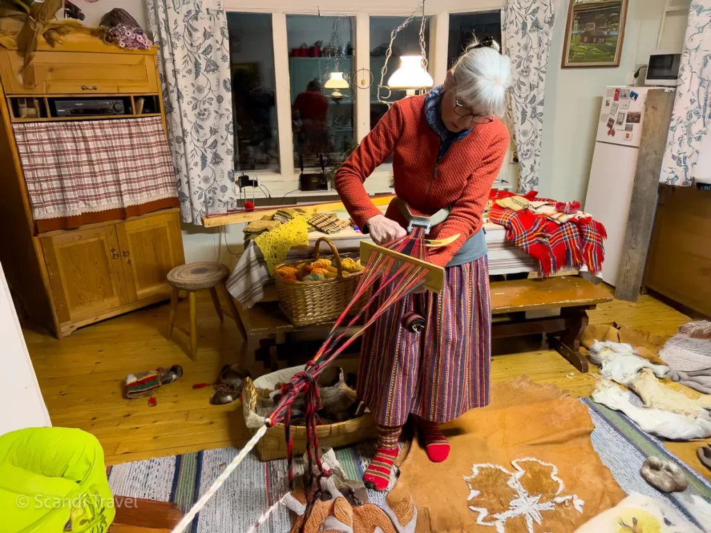 Woman weaving colorful woolen bands in a traditional Sami-style cabin, surrounded by rustic decor, during a Northern Lights tour in Lapland.