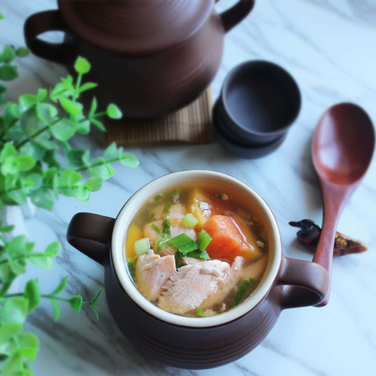 A bowl of Finnish Lohikeitto, a creamy salmon soup with vegetables, served in a brown ceramic bowl on a marble table with a wooden spoon and greenery. Perfect for a Finland Northern Lights tour culinary experience.