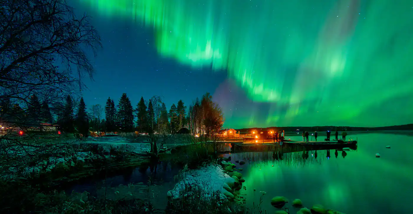 Northern Lights over a lake in Saariselkä, Finnish Lapland, with visitors watching the Aurora from a wooden pier near cozy cabins