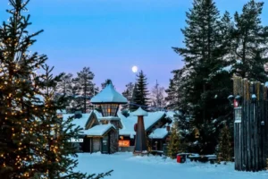 Snow-covered wooden village in Finnish Lapland at dusk, with a full moon rising above the trees and Christmas lights strung through the pines